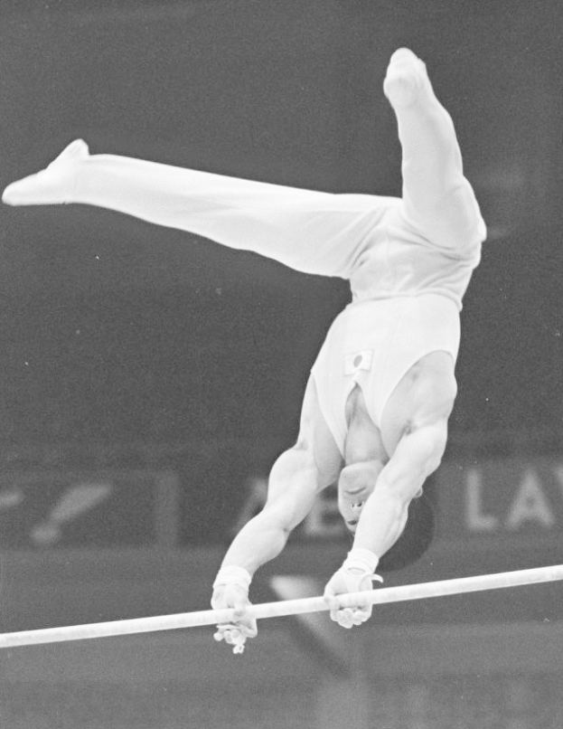 Ein Mann in einem Einteiler und Shorts führt Turnübungen an den parallelen Stangen aus, in der Luft mit ausgestreckten Armen und Beinen, Körper gebogen in einem anmutigen Bogen, Gesicht konzentriert und entschlossen, in einem Schwarz-Weiß-Foto mit einem unscharfen Hintergrund.