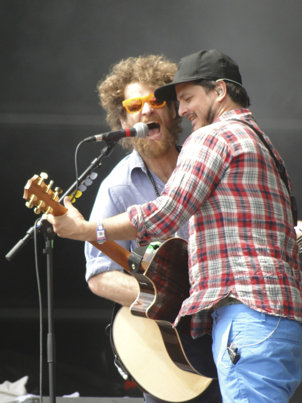 Zwei Männer auf der Bühne, einer in rot-blau kariertem Hemd mit blauen Hosen, der andere mit Brille und schwarzer Mütze, beide singen in Mikrofone und spielen Gitarren.