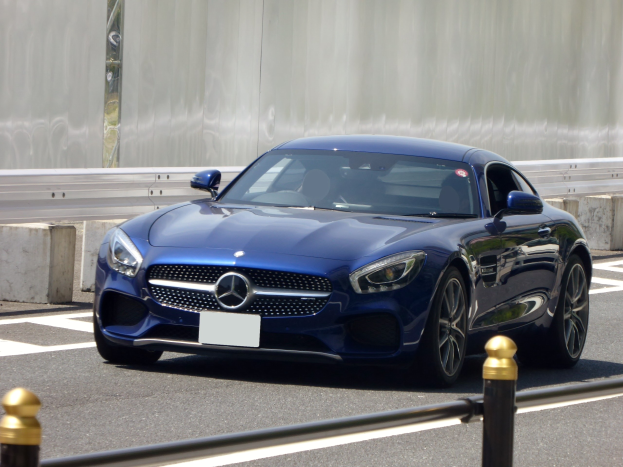 Blauer Mercedes-Benz AMG GT Coupé auf einer Straße fahrend, mit einer Wand im Hintergrund; schlank, aerodynamischer Design mit eckigen Scheinwerfern, großer grille, glänzenden Rädern und einem luxuriösen Lederinnenraum.