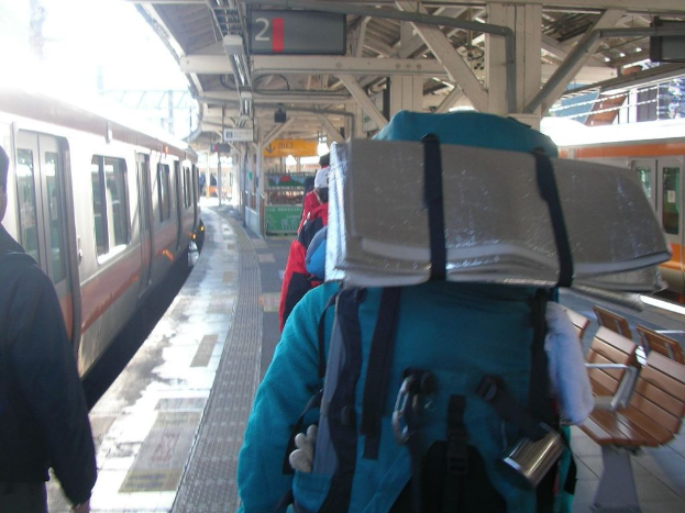 Ein Bahnhof mit Menschen neben einem Zug auf der linken Seite, einem Stand in der Mitte, einem weiteren Zug auf der rechten Seite und einer Bank in der rechten unteren Ecke.