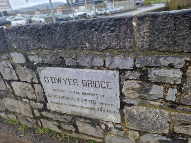 Ein Steinwall mit einer Tafel, die "O'dwyer Bridge" trägt, vor einer Straße mit Fahrzeugen, Polen, Bäumen, Gebäuden und einem klaren blauen Himmel.