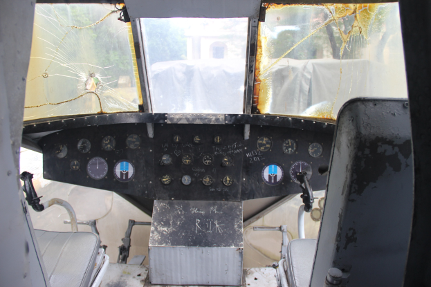 Cockpit-Inneneinrichtung eines Flugzeugs mit einer zersplitterten Frontscheibe, zwei Pilotensitzen und mehreren Steuerinstrumenten, die eine Äußere Ansicht von Bäumen und einem Gebäude durch die zerbrochene Scheibe zeigen.