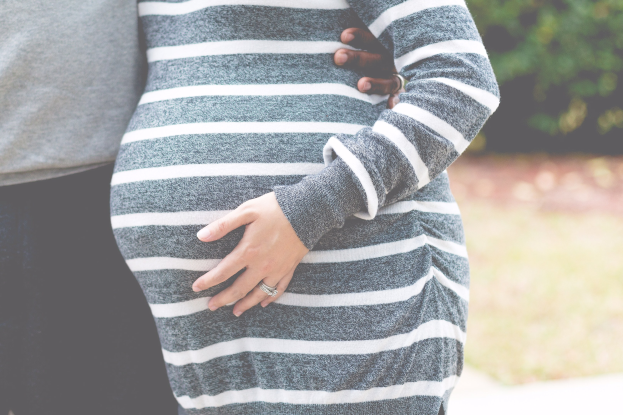 Schwangere Paare nebeneinanderstehend mit Händen auf dem Bauch, tragend Maternity-Kleidung, umgeben von Bäumen.