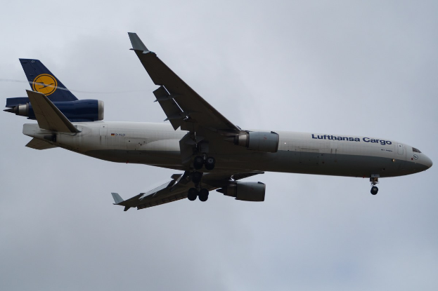 Ein Lufthansa Cargo Boeing 777-300ER im Flug, mit ausgefahrenen Tragflächen und aktivierten Triebwerken, mit sichtbarem Text auf der Rückseite gegen einen wolkenfüllten Himmel.