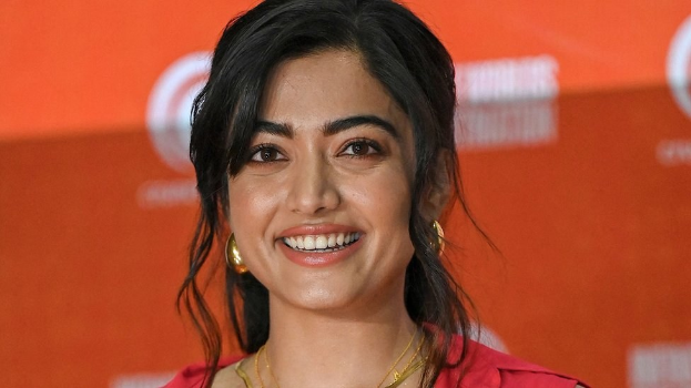 Eine Frau in einem roten Kleid mit einem eleganten Dutt und dezentem Make-up steht vor der Kamera und lächelt, trägt schwarze High Heels und hat ein hellgelbes Banner mit der Aufschrift "Sonam Kapoors Beste Schönheits Looks vom Cannes Film Festival" im Hintergrund.