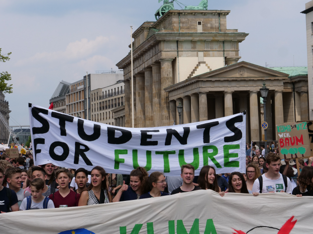 Gruppe von Schülern marschiert in Berlin mit einer leuchtend bunten 'Fridays for Future'-Schultasche vor einem Hintergrund aus Gebäuden, Bäumen und Himmel.
