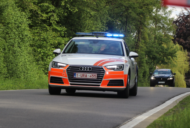 Polizeiauto (Audi A4) fährt auf einer grasbewachsenen Straße mit zwei Insassen, umgeben von Bäumen unter einem klaren blauen Himmel.