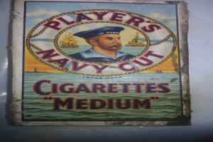 Plakat mit einer Person in einer marineblauen Uniform mit weißem Hemd und roter Krawatte, die Arme verschränkt und einen strengen Ausdruck hat, wirbt für "Player's Navy Cut Cigarettes Medium" in fetter schwarzer Schrift innerhalb eines weißen Randes.
