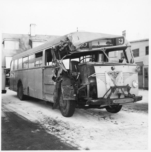 Ein Schwarz-Weiß-Foto eines schwer beschädigten Busses am Straßenrand mit eingedellter Front und zersplitterten Fenstern und Gebäuden im Hintergrund.