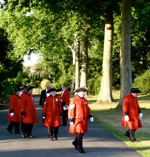 Eine Gruppe älterer Männer in einheitlicher Kleidung, mit einem Mann in Hemd, Blazer, Hose und Schuhen, der auf einer Straße steht, mit Gebäuden, Bäumen, Pflanzen und Gras im Hintergrund.