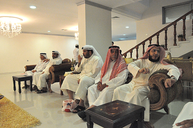 Männer sitzen auf einer Couch mit einer Treppe und einem Kronleuchter im Hintergrund.
