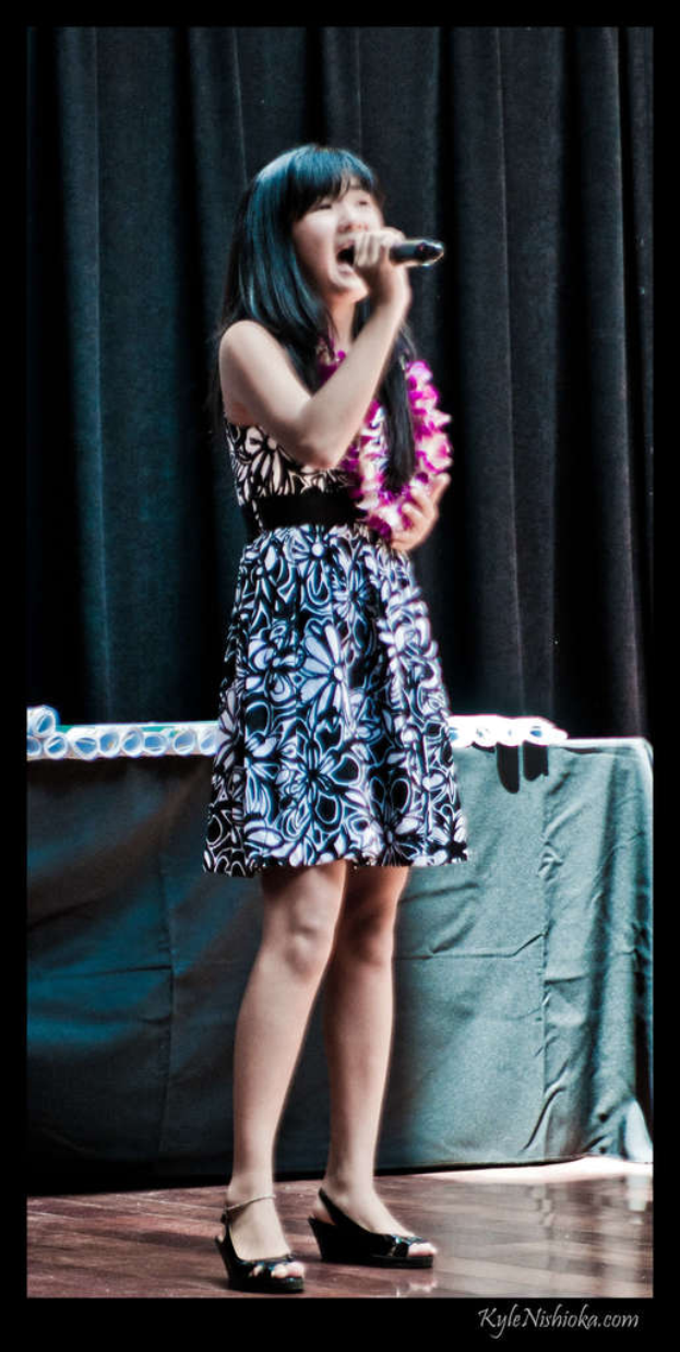 Eine Frau in einem bunten Kleid singt auf der B├╝hne in ein Mikrofon, mit einem Tisch mit Papieren und einem Vorhang im Hintergrund.