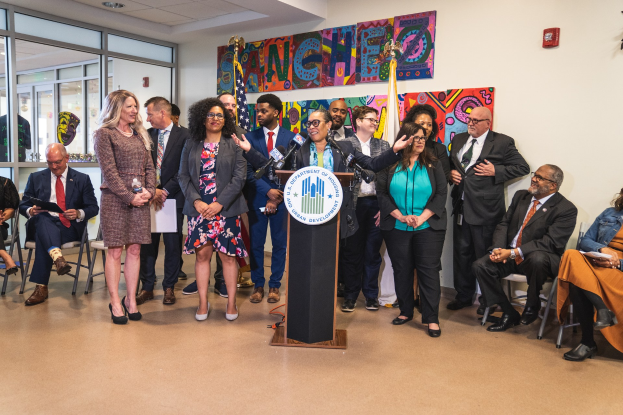 Gruppe von Menschen vor einem Rednerpult mit Mikrofon und einer Tafel mit Text 'Sancho-Stadtrat tritt dem Stadtrat bei', mit sitzenden und stehenden Teilnehmern, Flaggen, Feuerlöscher und Deckenleuchten im Hintergrund.