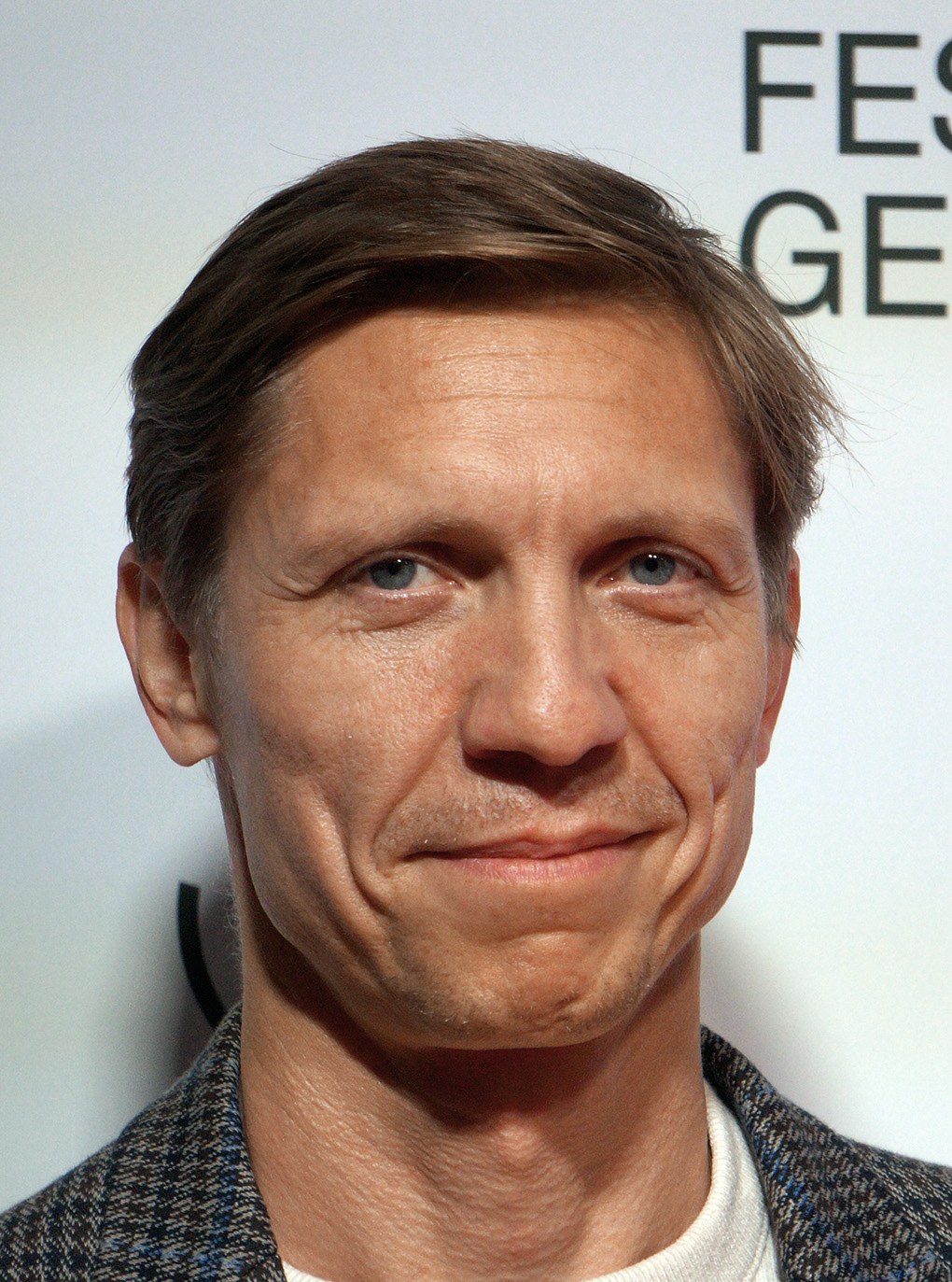 Ein Mann im Anzug und Krawatte lächelt in die Kamera, im Hintergrund ist eine Tafel mit der Aufschrift "Eddie Redmayne Vermögen" zu sehen.