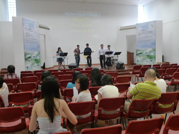 Eine Bühnenmitte mit zwei Gitarristen und anderen Sängern und Musikern in den Ecken, mit Bannern neben der Bühne und einem Publikum davor mit Papieren und Büchern.