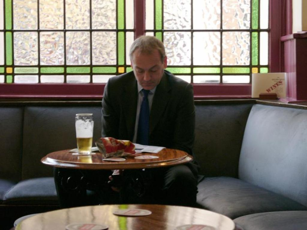 Ein Mann sitzt auf einer Couch mit Papieren und einem Glas auf einem Tisch davor, ein Fenster im Hintergrund.