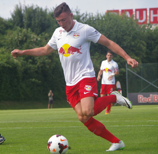 Ein Mann in einem Fussballtrikot schießt einen Ball auf einem grünen Feld mit Bäumen und einem klaren blauen Himmel im Hintergrund, wobei ein Schild mit der Aufschrift "RB Leipzig" zu sehen ist.