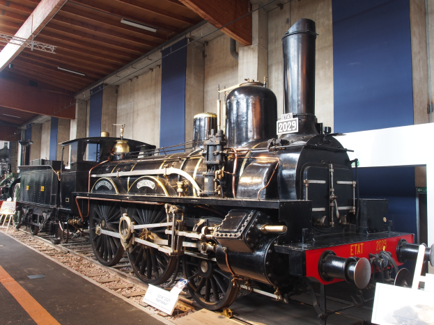 Alte Dampflokomotive in einem Museum ausgestellt, umgeben von Eisenbahnschienen, Informationsschildern und anderen Ausstellungsstücken, mit einer Wand und beleuchteter Decke im Hintergrund.