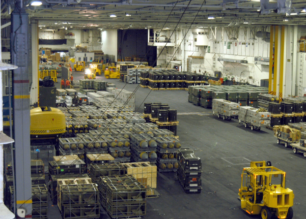 Großer Lagerraum mit Lkw, Containern, Menschen, Deckenbeleuchtung, Metallstangen und einem sichtbaren Rad.