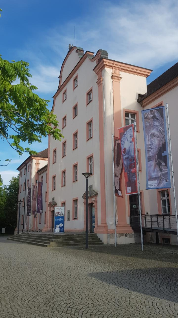 Ein großes Gebäude mit zahlreichen Fenstern, umgeben von Straßenmasten, Straßenlaternen, Werbetafeln, Treppen, Geländern, Bäumen und einem bewölkten Himmel, identifiziert als das Museum für Geschichte und Kultur von Baden-Württemberg.