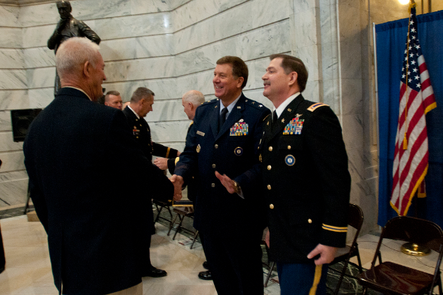 Menschen schütteln Hände mit Stühlen dahinter, Statue neben einer Wand im Hintergrund und Flagge und Vorhang rechts.