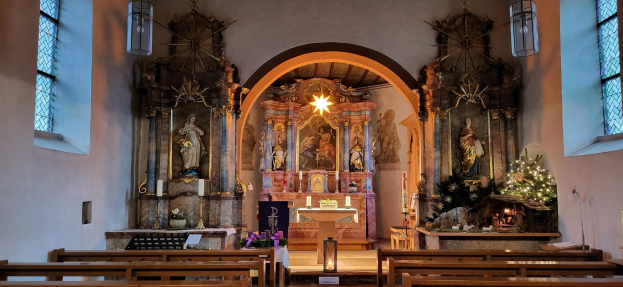 Innere einer Kirche mit hölzernen Kirchenbänken, Kerzenständern, Blumenbouquets, Statuen, Lichtern, Fenstern und einem geschmückten Weihnachtsbaum im Hintergrund nahe einem Bogen.