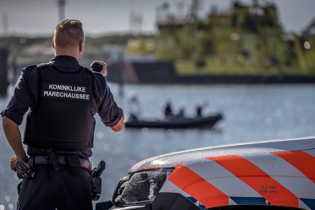 Ein Polizist in Uniform steht neben einem Polizeiwagen in der Nähe eines Gewässers und hält ein Funkgerät in der Hand, im Hintergrund ist ein Boot mit ein paar Menschen auf dem Wasser unter einem klaren Himmel zu sehen.