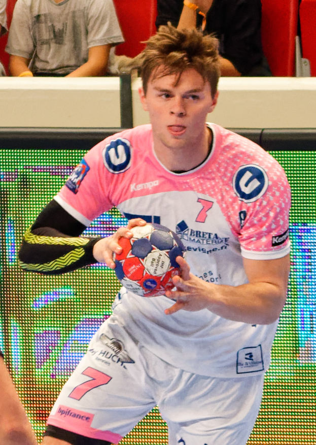 Ein Mann in einem pinken Hemd mit einem Fußball, daneben eine weitere Person mit der Hand im Bild, während im Hintergrund Menschen auf Stühlen sitzen.