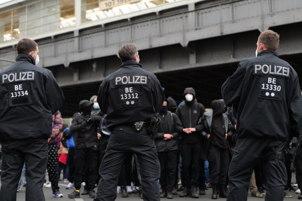 Eine Gruppe von Polizeibeamten in schwarzen Uniformen und Masken steht vor einer Menge, mit einer Brücke und einem Gebäude im Hintergrund, während einer Demonstration in einer Stadt.