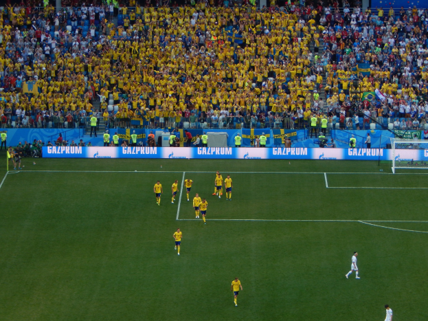 Gruppe von Menschen, die Fußball auf einem Rasenfeld mit zwei Toren spielen, beobachtet von einer großen Menge in Sitzplätzen mit Tafeln im Hintergrund.