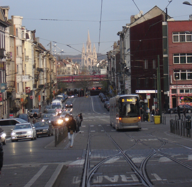 Eine belebte Stadtstraße mit mehreren Fahrzeugen, Fußgängern auf dem Gehweg und Gebäuden auf beiden Seiten, mit einer Tram, einer fernen Brücke, Bäumen und einem klaren blauen Himmel im Hintergrund.