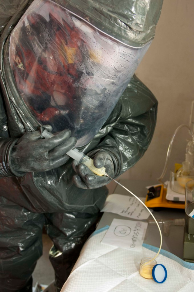 Eine Person in einem Laborkittel, Helm und Handschuhen injiziert eine Substanz in eine Flasche auf einem Tisch, auf dem auch Papiere und eine Waage liegen.