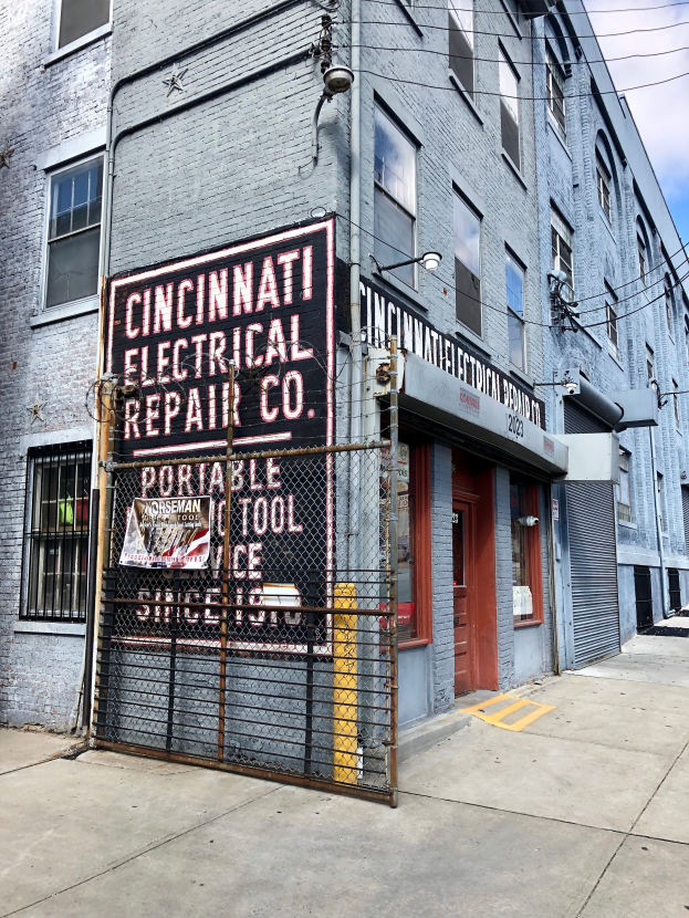 Das Gebäude der Cincinnati Electrical Repair Co. in Brooklyn, New York City, mit Fenstern, einer Tür, einem Schild, einem Metallzaun, einem Straßenmast und Drähten unter einem bewölkten Himmel.
