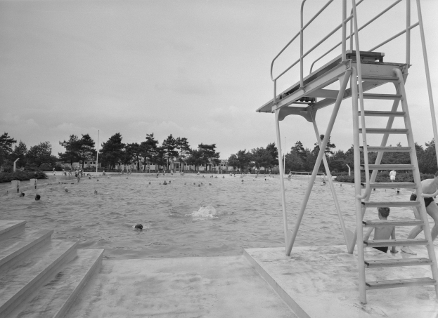 Schwarz-weiß-Foto von Menschen, die im Wasser an einem Strand schwimmen, mit einem Rettungsturm auf der rechten Seite, einer Treppe, die hinaufführt, Bäumen und Pfählen im Hintergrund und einem klaren Himmel.