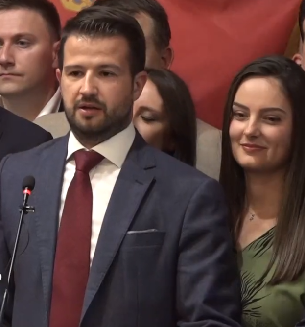 Ein Mann im Anzug und Krawatte steht an einem Podium mit einem Mikrofon und spricht zu einer Menge mit einem Banner im Hintergrund.