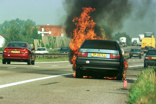 Ein Auto in Flammen auf dem Seitenstreifen, umgeben von anderen Fahrzeugen, mit Bäumen, Gebäuden und einem klaren blauen Himmel im Hintergrund und einem Feuerlöscher auf der rechten Seite.
