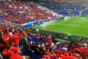 Ein volles Stadion mit Sitzplätzen und Stehplätzen, Fahnen, ein Torpfosten und Deckenbeleuchtung, während die Menge gespannt auf den Beginn eines Fußballspiels wartet.